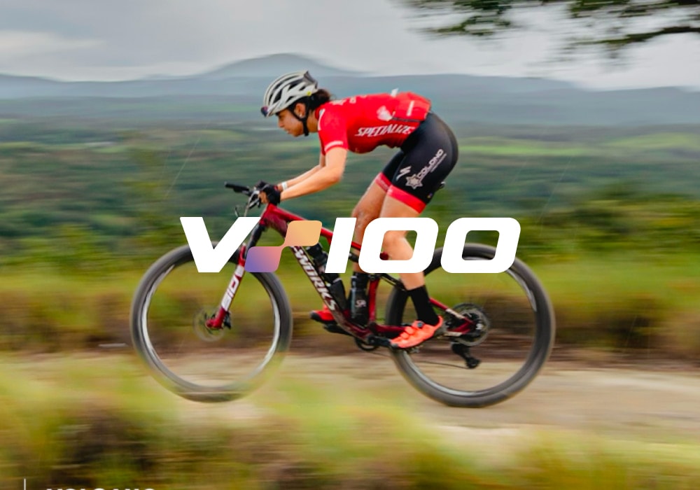 A mountain biker in a red Specialized kit, motion-blurred at speed, with the V100 logo across the middle of the frame